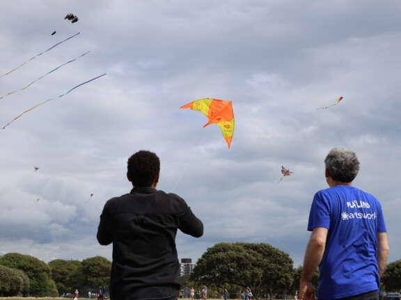 young and older person looking away from the camera, young person is flying an orange and yellow arrow shaped kite that says 'playland' on it in a cloudy sky, other kites are visible