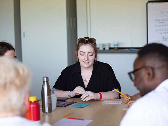 An Artswork staff member looking down and smiling at their drawing, surrounded by fellow team members. She has brown hair, sunglasses on top of their head, and is wearing a black dress.
