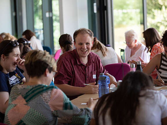 A group of Artswork staff members sat at a table, one is talking and smiling, the others are listening whilst they use pencils to sketch mini self-portraits.