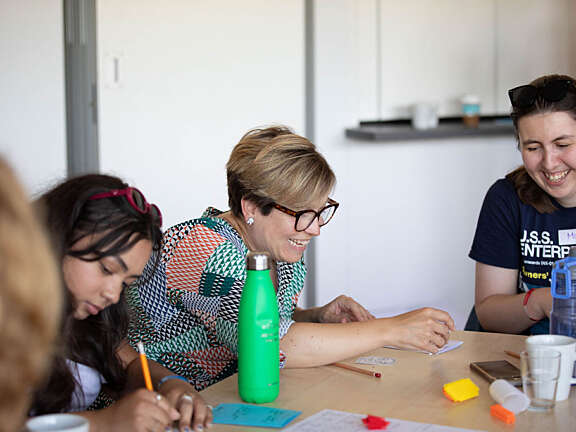 Three Artswork members taking part in a creative activity, the young person on the left, Rhe, is concentrating intently on her artwork, and the woman in the middle, Beccy, is laughing with Mara, another young person on her right