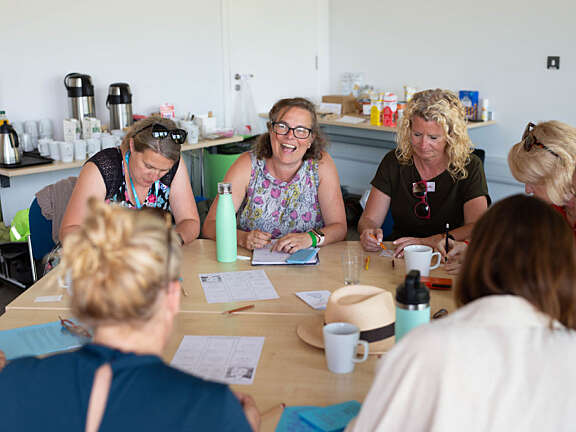 A group of Artswork staff members sat at a table talking and smiling, whilst they use pencils to sketch mini self-portraits.