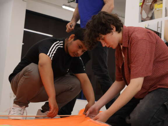 two young people crouched down holding orange fabric and a dowel to make a kite