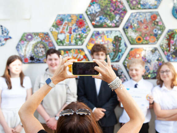 An artist using their smartphone to take a photo of a group of young people in front of a mosaic mural they designed and helped create