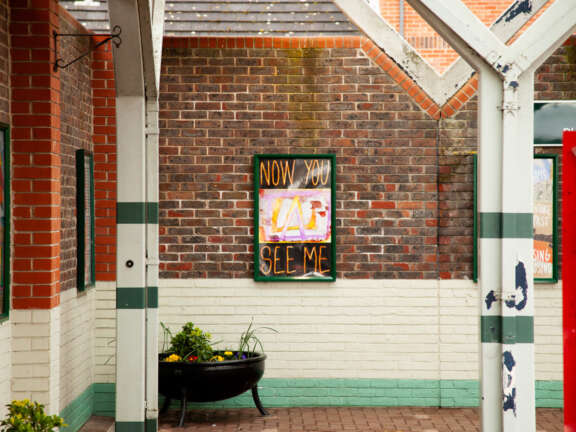 Young person's artwork displayed on the wall of a local train station, with graphic text that says "now you see me"