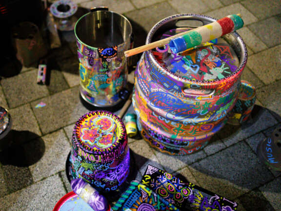 A pile of instruments made from recycled materials as part of a 'Junk Band'. The surfaces of drums, shakers, and cymbals have been covered in colourful doodles by young people.