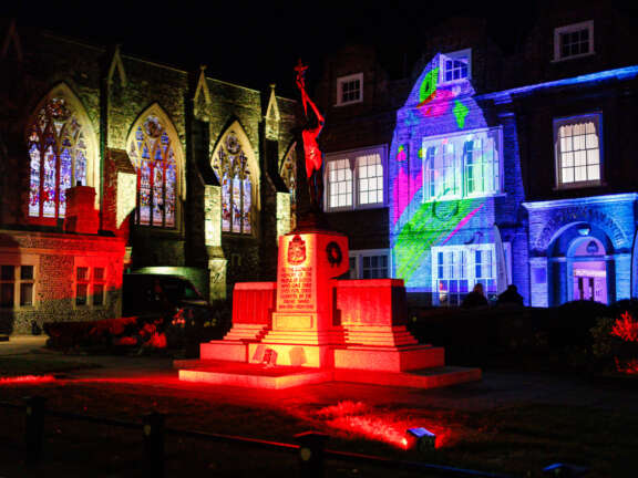 A building that has been illuminated with multicoloured projections of young people's artworks, a bright red light is shining on a statue in the foreground