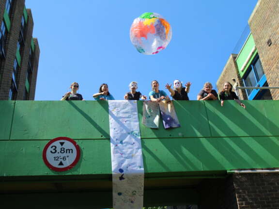7 young people stand on a bridge with blue sky behind them. They all look happy and throw a colourful paper globe off the bridge.
