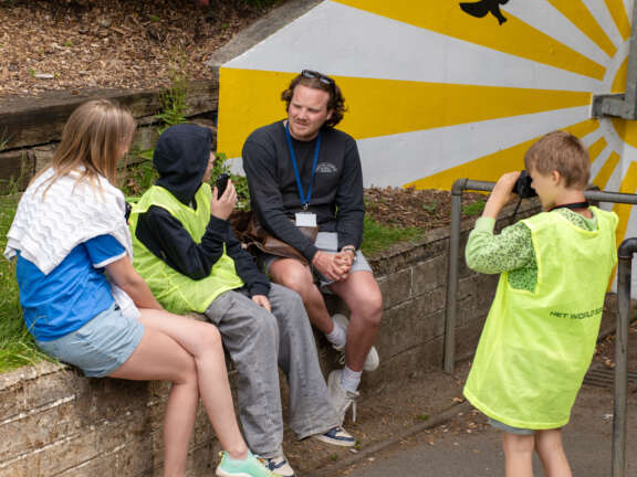 a man being interviewed by a young boy, with another young boy filming and a woman sat listening, all in front of a ray of sun mural