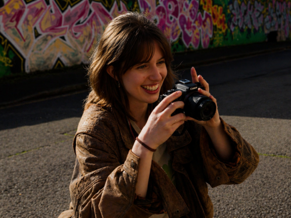 A young woman smiles outside on the street as she holds up a camera. A colourful mural is behind her.