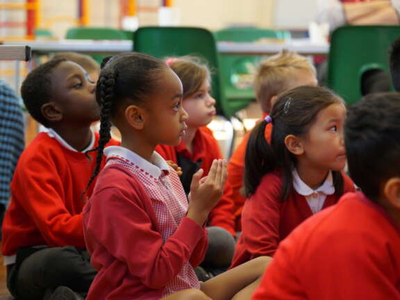 A group of young primary school children sit on the floor looking ahead to someone speaking.