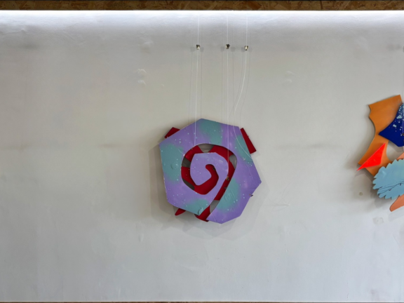Three abstract, colourful wall sculptures hanging against a gallery wall. They resemble the shape of a flower, spiral, and face