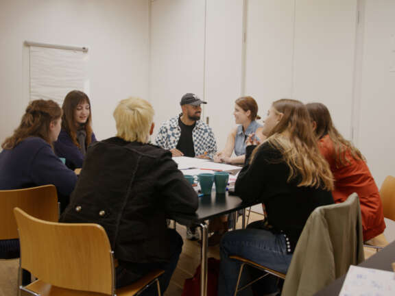 Princess Eugenie sits at a table with a small group of young people and appears in discussion with a young man to the left.