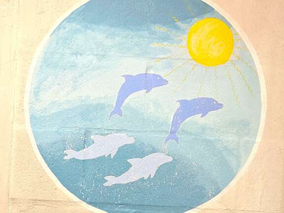A circular mural in pastel colours of dolphins swimming and jumping with a sun, painted onto a plain wall