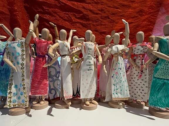 about 15 small wooden mannequins arranged into a group photo style pose all wearing fabric tied into dresses and decorated with fabric trims and pens