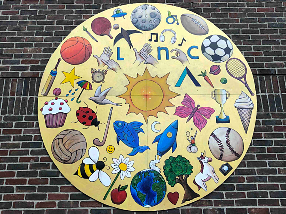 Circular golden artwork decorated with painted pictures including a sun, butterfly, basketball, hands spelling out BSL letters and more. On wall.
