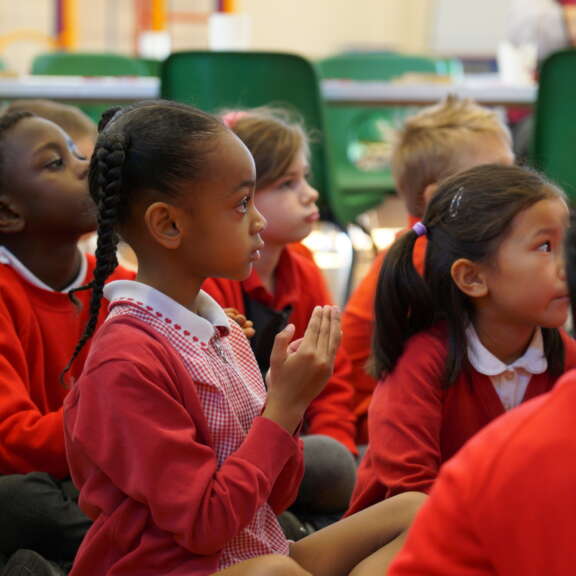A group of young primary school children sit on the floor looking ahead to someone speaking.