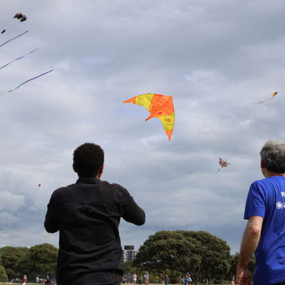 young and older person looking away from the camera, young person is flying an orange and yellow arrow shaped kite that says 'playland' on it in a cloudy sky, other kites are visible