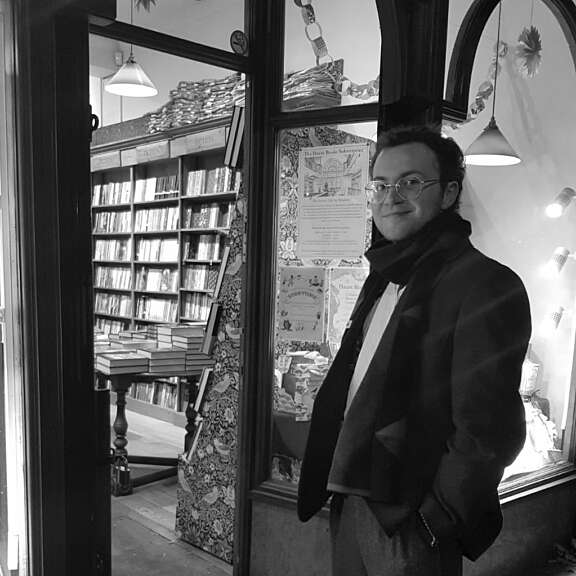 man with glasses and a long scarf standing in front of a book shop