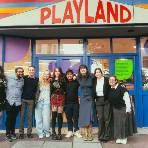 9 young people (under 30) stand outside Playland with the sign in the background - the people are a mix of genders and ethnicities.