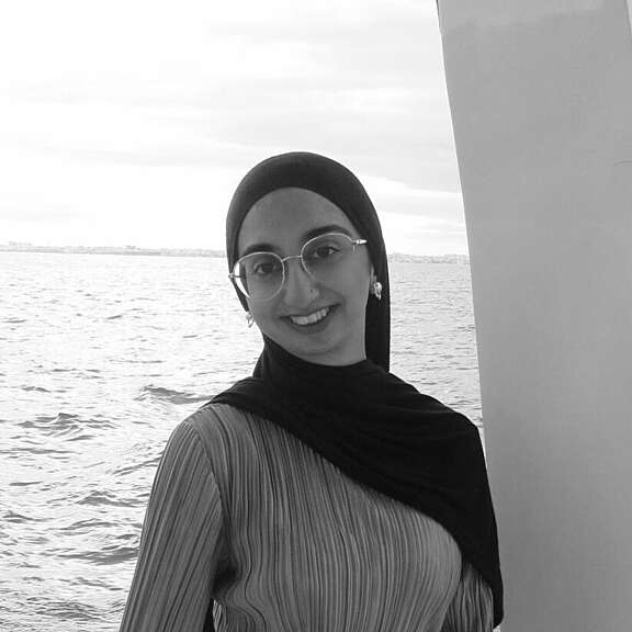 Sanna a young woman smiles at the camera, wearing a hijab and glasses. the sea is visible behind her