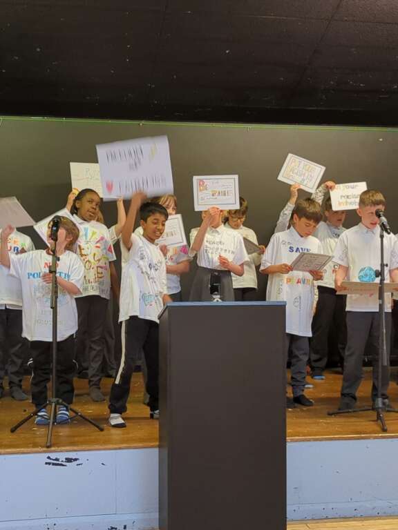 Year 5 pupils on stage wearing decorated t-shirts and holding signs with messages on them, in performance