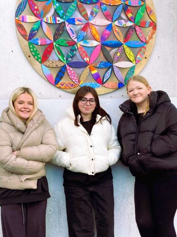 three young girls standing in front of a flower of life geometric artwork hung on wall made from many individually decorated petals
