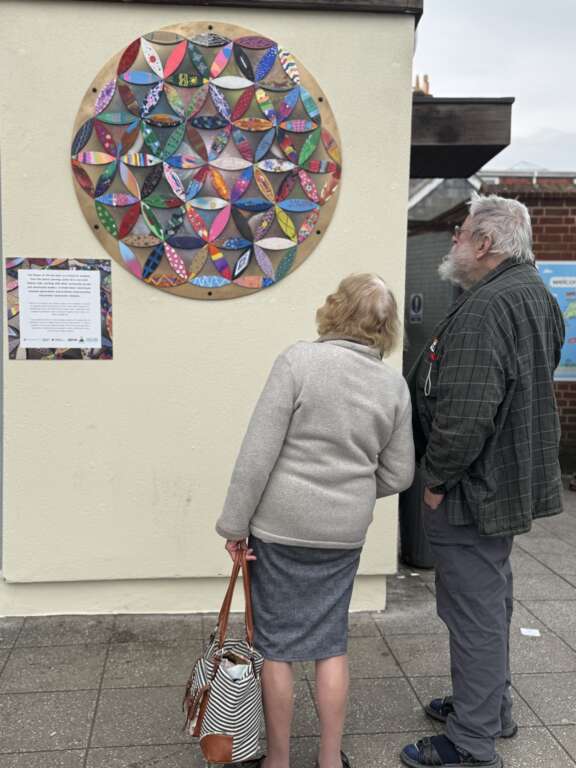 two elderly people looking at a flower of life geometric artwork on wall made from many individually decorated petals