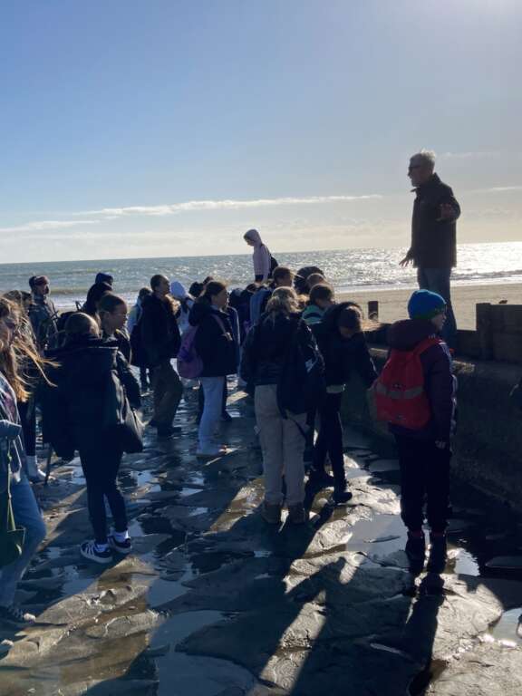a group of children gather on a beach in the winter sun, facing a man standing on a wall addressing them