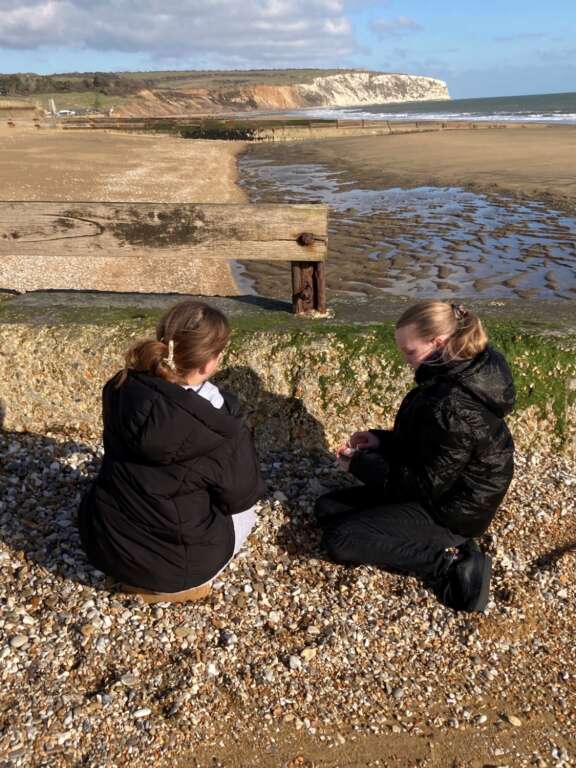 Two girls in coats sat on beach, appear to be looking at something