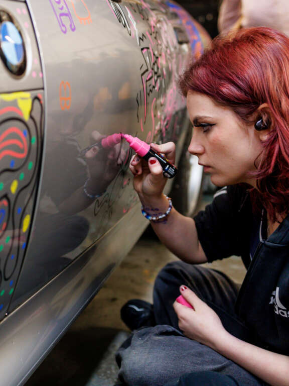 A young college student drawing directly onto a donated silver car using acrylic paint pens that is being transformed into a colourful 3D mural