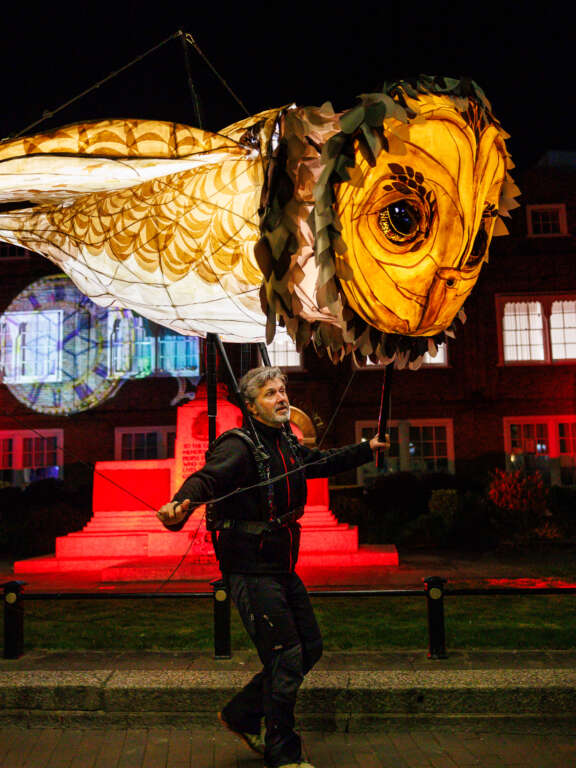 night scene with a large glowing willow lantern in the shape of a flying owl, operated by a man underneath