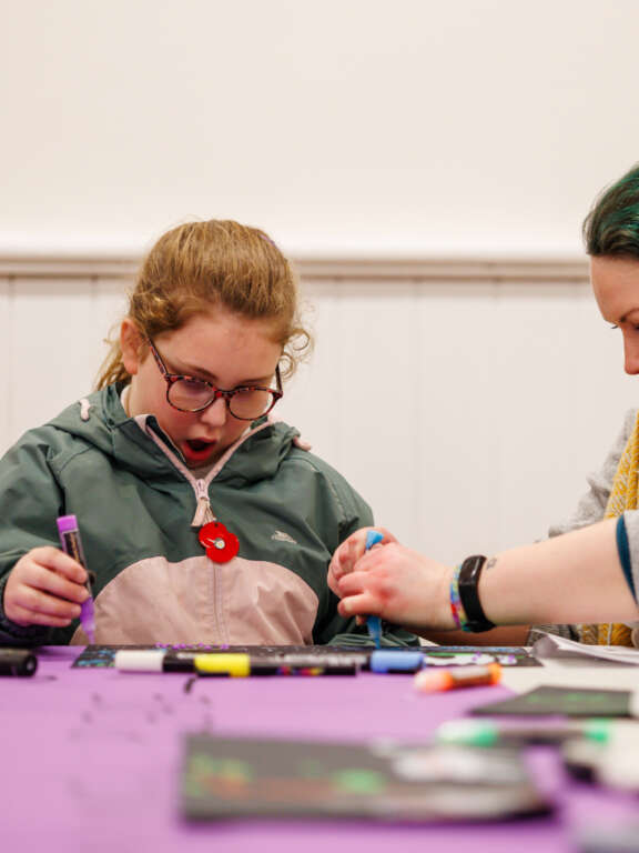 A young person sat at a table next to an adult, participating in a creative workshop using acrylic paint pens