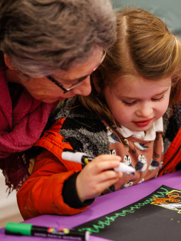 A young girl drawing using acrylic paint pens alongside her grandmother