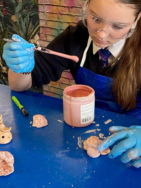 secondary age girl in school uniform wearing goggles and protective gloves holds a paintbrush with peach coloured ceramic glaze for a sculpture she has made