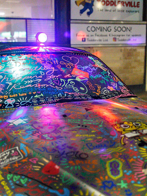 A silver car that has been completely covered in various colourful drawings, doodles, and text by young people. The car is illuminated by a purple light, shining through the windows and highlighting the drawings on the windscreen