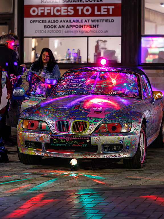 A crowd gather around a silver car that has been completely covered in various colourful drawings, doodles, and text by young people.