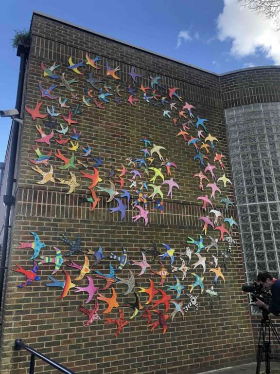 Flock of brightly coloured ceramic swallows arranged in a large spiral flight pattern, spanning multi story wall
