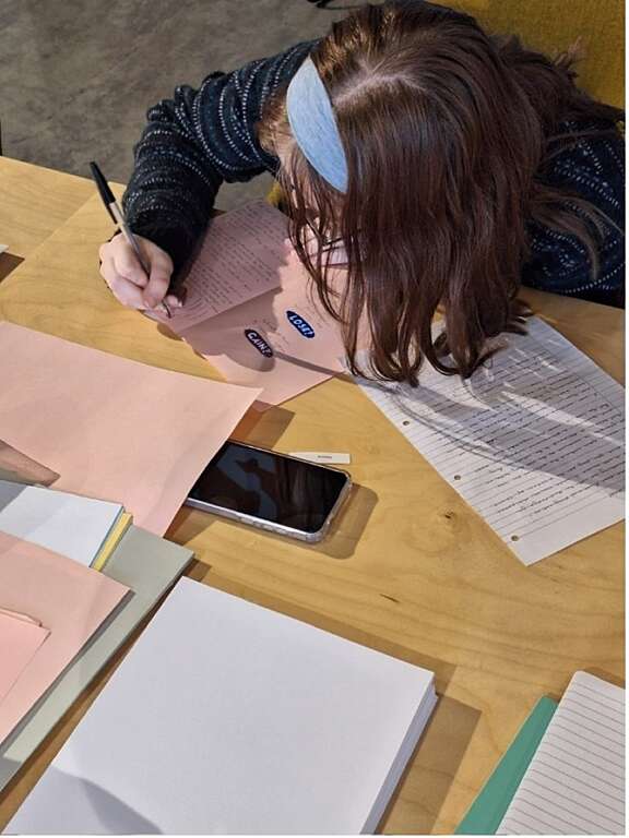 Picture taken from above of person with red hair and a blue headband handwriting a poem on pink paper