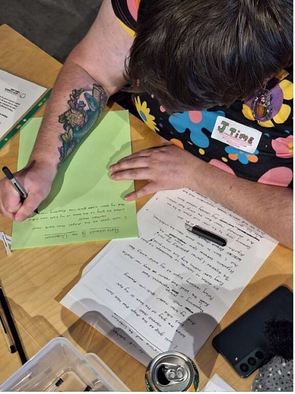 Picture taken from above of person with tattoo on arm handwriting a poem on green paper