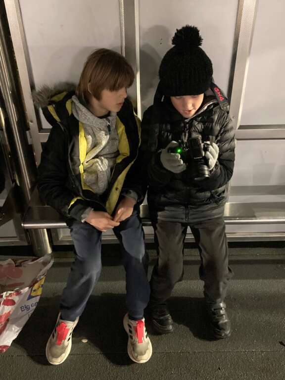 two young boys sat on train station bench looking at a camera, seemingly in discussion