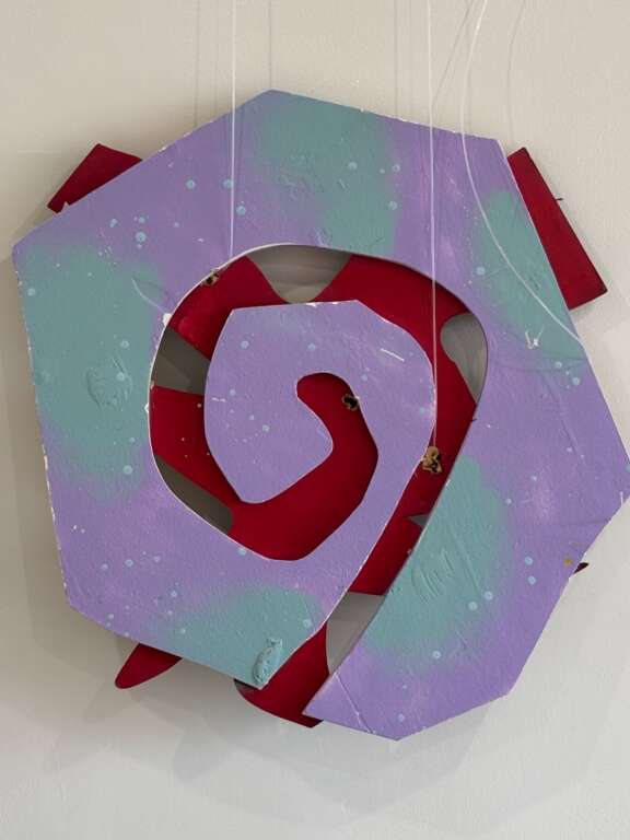 An abstract, colourful wall sculpture hanging against a gallery wall. It resembles the shape of a spiral and combines lilac and teal colours.