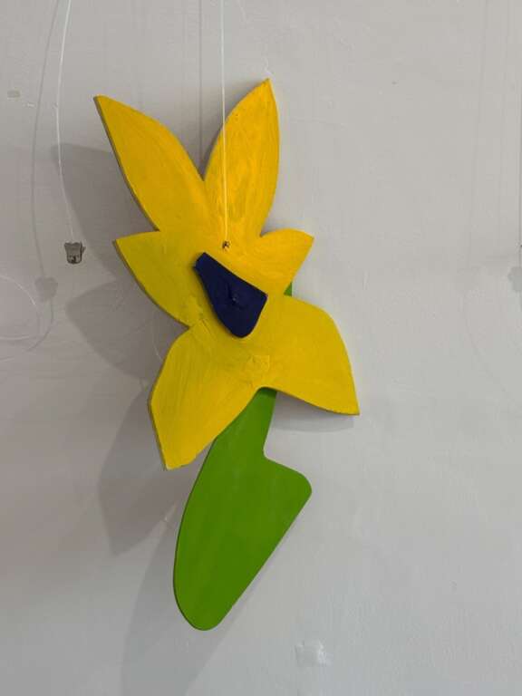 An abstract, colourful wall sculpture hanging against a gallery wall resembling a bright yellow Daffodil.