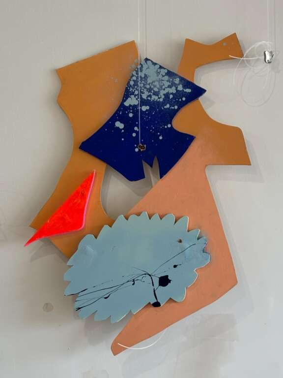 An abstract, colourful wall sculpture hanging against a gallery wall. It resembles the shape of a face, complete with a bright orange triangle nose.