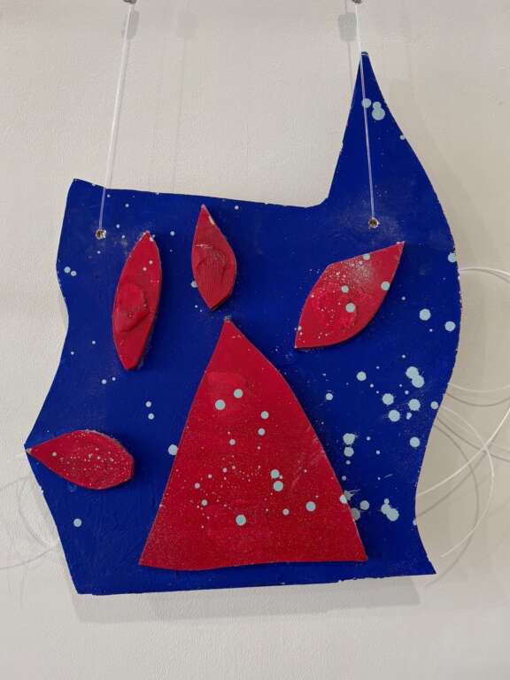 An abstract, colourful wall sculpture hanging against a gallery wall. It resembles the shape of a red pawprint against a blue background.