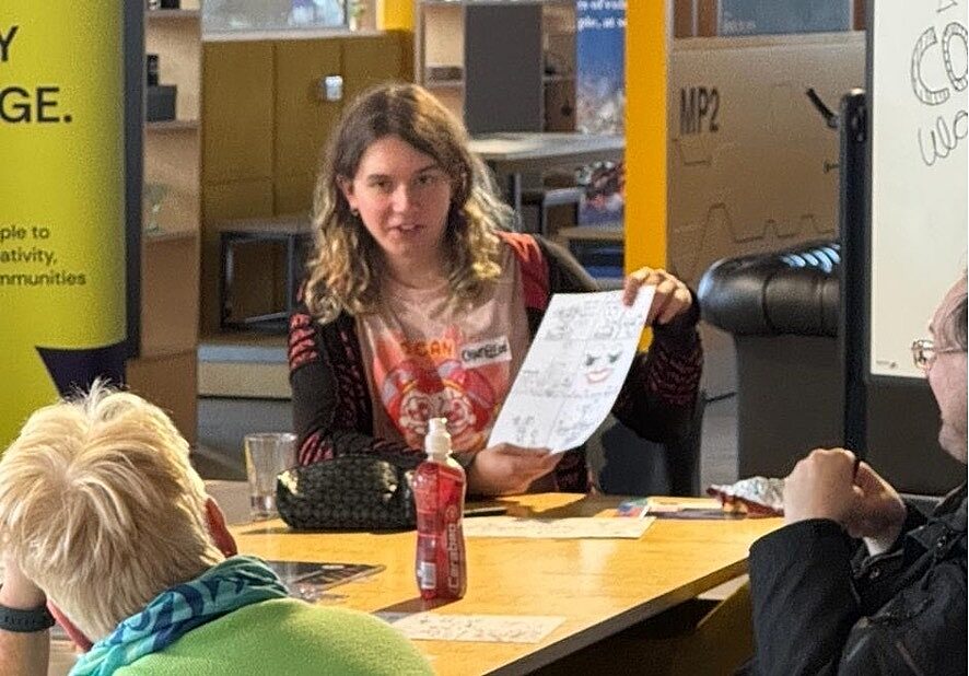 young person holding paper with hand drawn cartoon on it, appears to be explaining it to others around the table