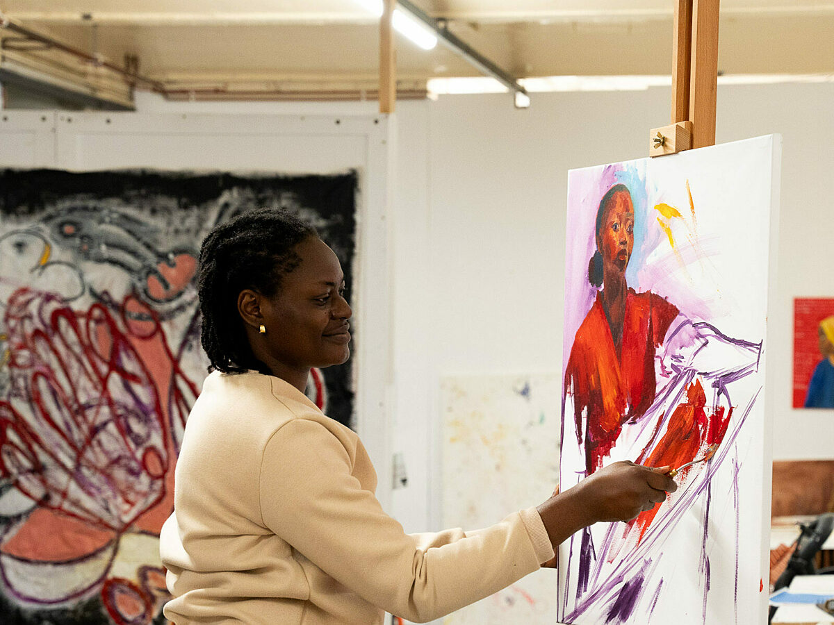 An artist in her studio at BACKLIT Gallery. The artist is a young black woman. She is wearing a light yellow top with long sleeves, and blue jeans. She stands side-on to the camera and is painting a portrait on an easel. The portrait is of a black woman with a red top. The background of the painting is white and incomplete. The artist's brush touches the canvas, mid-action.
