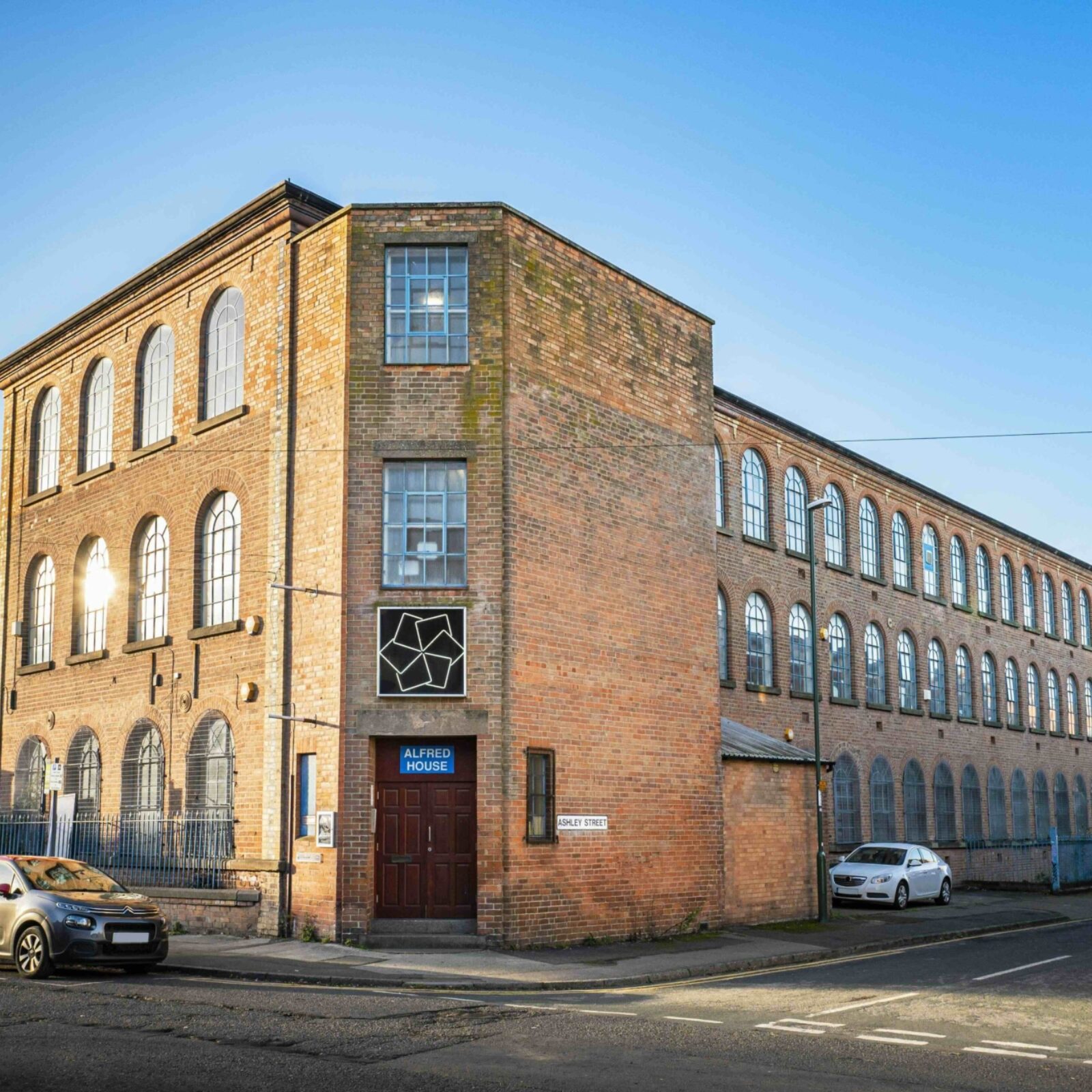 Exterior of BACKLIT Gallery, a large red-brick Victorian factory building with tall arched windows on a street corner in Nottingham. The bright blue sky hugs the building.