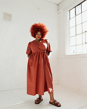 Portrait of Kim Thompson standing in a bright, minimal studio space, wearing a loose muted orange wrap dress and sandals, with a large red afro hairstyle, hands relaxed behind her back, and sunlight streaming in through a window to her right.