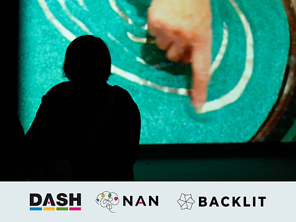 Silhouetted visitor watching a projected close-up of a hand drawing curved white lines in sand. DASH, NAN and BACKLIT logos appear below.