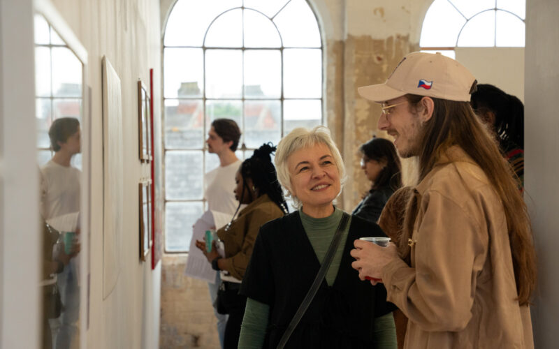 Smiling people talking at an exhibition inside BACKLIT Gallery, with large arched windows, white walls, and framed artworks on display.